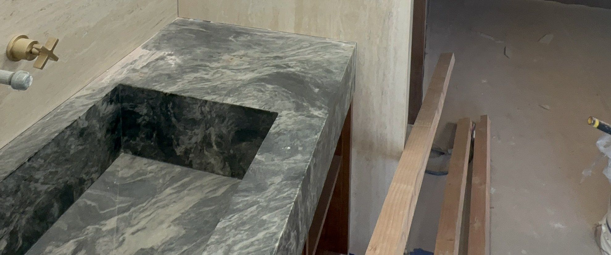 A green veined stone vanity with an integral sink against travertine walls, in a primary bathroom under construction in an Akopi Builders custom home in Bel Air