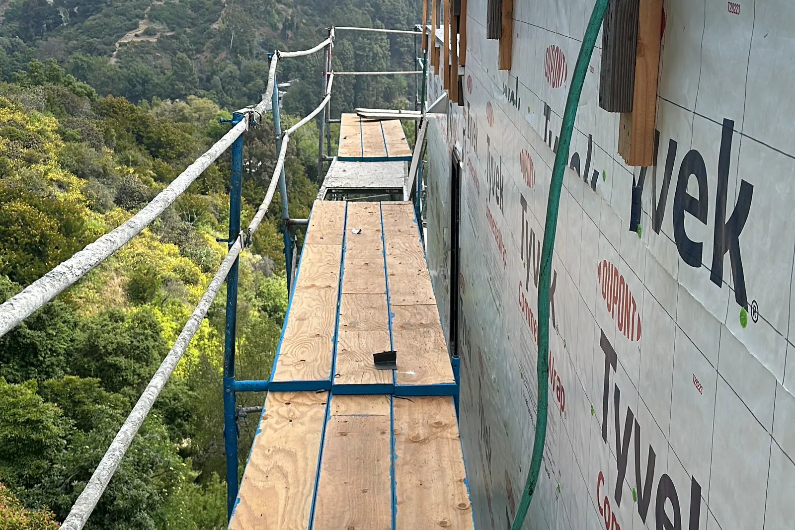 Scaffolding planks along a house-wrapped wall on a hillside home under construction in Bel Air, with trees and brush on the slopes below, on an Akopi Builders project.