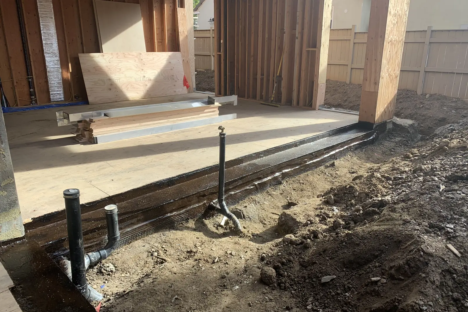 The slab edge of a house under construction with waterproofing along the footing and black drain pipes stubbed up before backfill, on an Akopi Builders project.