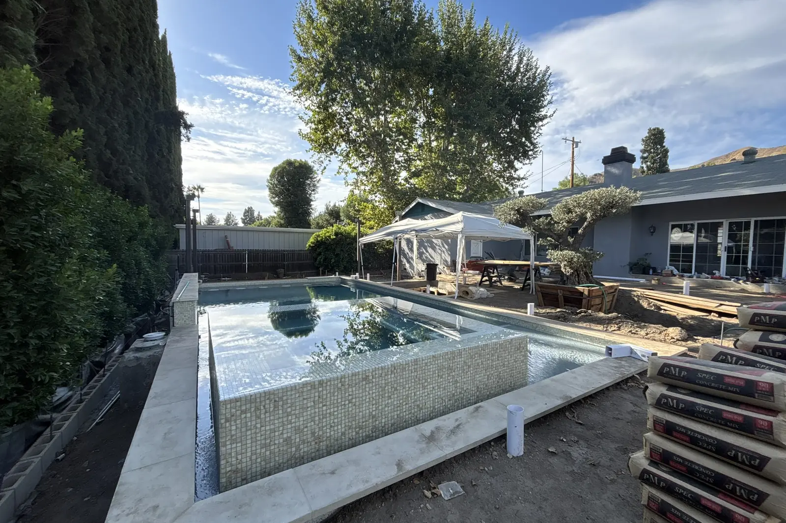 A new pool with pale stone coping and a mosaic tiled spa wall, the deck area still bare, at Arnwood, an Akopi Builders backyard build in Los Angeles.