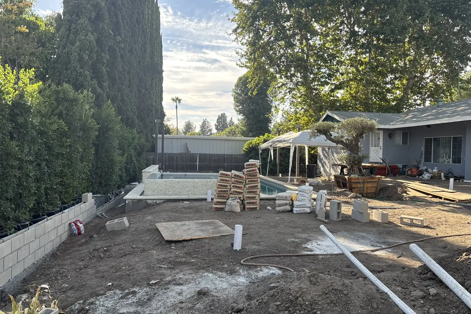 Backyard during site work with new utility pipes in the ground and paving materials stacked by the pool, at Arnwood, an Akopi Builders build in Los Angeles.