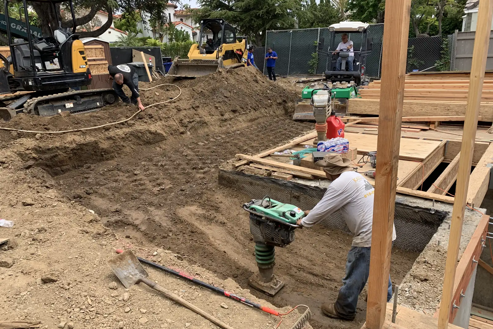 A residential lot with a small excavator, fresh foundation forms and a worker compacting soil, on an Akopi Builders project in Santa Monica.
